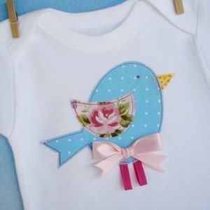 May include: A white baby onesie with a blue and white polka dot bird applique. The bird has a pink rose and a pink bow.