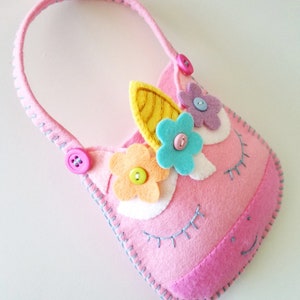 Puede incluir: Cartera de unicornio de fieltro rosa con un cuerno amarillo, ojos blancos y flores de fieltro de colores. La cartera tiene un bot&oacute;n rosa y un bot&oacute;n azul.