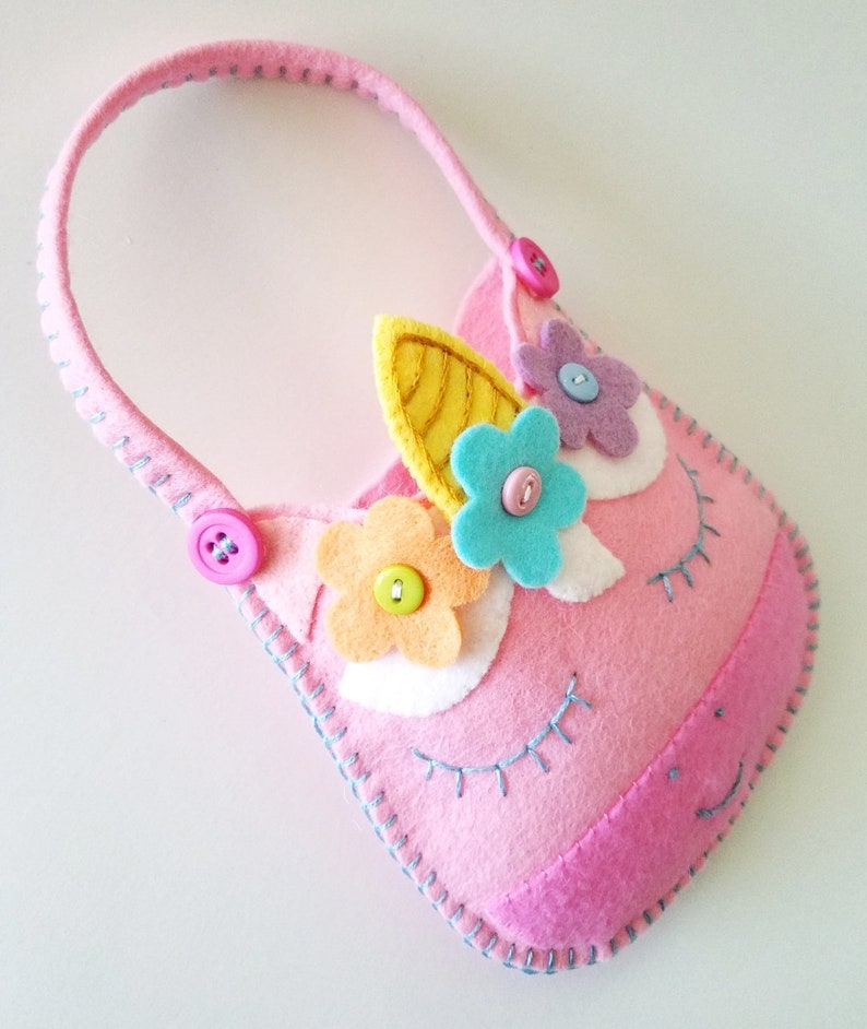 Puede incluir: Cartera de unicornio de fieltro rosa con un cuerno amarillo, ojos blancos y flores de fieltro de colores. La cartera tiene un bot&oacute;n rosa y un bot&oacute;n azul.