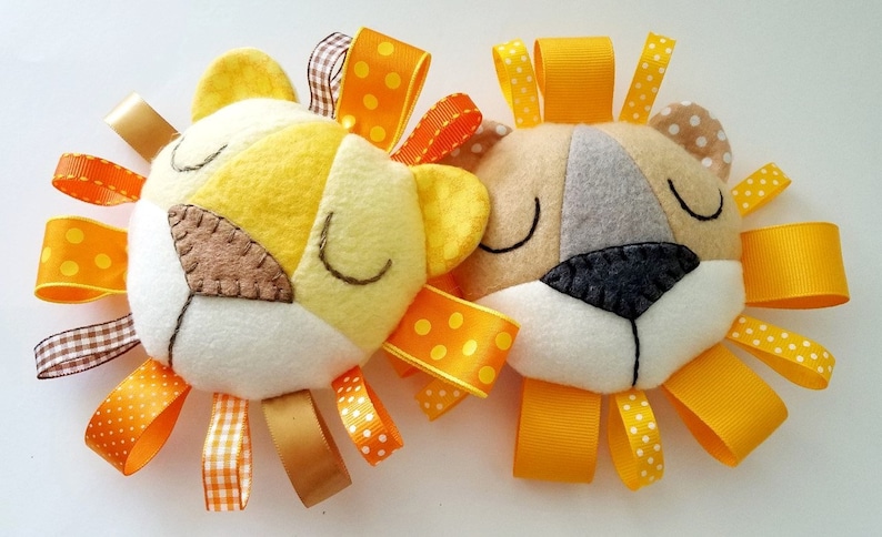 Op de afbeelding: Twee pluche speelgoedjes in de vorm van een leeuwenkop met gele en oranje lintstaarten. De leeuwen hebben gesloten ogen en zijn gemaakt van vilt.