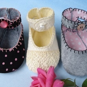 May include: Three pairs of felt baby shoes with different patterns. The first pair is black with pink polka dots, the second pair is white with a lace pattern, and the third pair is gray with a pink polka dot lining.