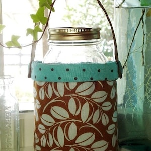 May include: A brown fabric jar holder with a teal polka dot trim and a white leaf pattern. The holder has a brown leather handle and is holding a clear glass jar with a gold lid.
