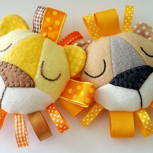 Op de afbeelding: Twee vilten baby speelgoedjes in de vorm van een leeuwenkop met gele, oranje en bruine linten. De leeuwen hebben gesloten ogen en zijn gemaakt van verschillende kleuren vilt.