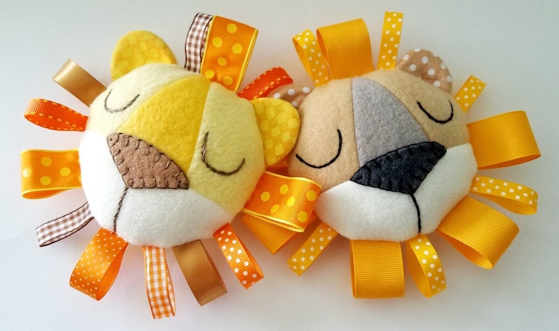 Op de afbeelding: Twee vilten baby speelgoedjes in de vorm van een leeuwenkop met gele, oranje en bruine linten. De leeuwen hebben gesloten ogen en zijn gemaakt van verschillende kleuren vilt.