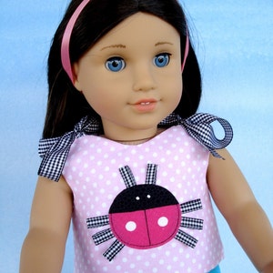 May include: A pink and white polka dot dress with a ladybug applique on the front. The dress has a blue and white floral print skirt. The doll is wearing a pink headband.