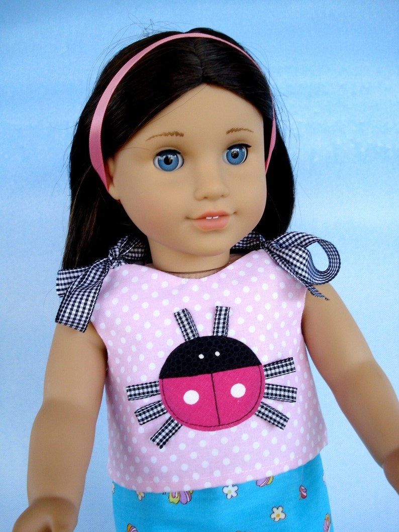 May include: A pink and white polka dot dress with a ladybug applique on the front. The dress has a blue and white floral print skirt. The doll is wearing a pink headband.
