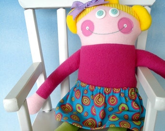 May include: A handmade doll with yellow hair, pink cheeks, and a bright pink sweater. The doll is wearing a blue and green skirt with a circle pattern and is sitting in a white rocking chair.