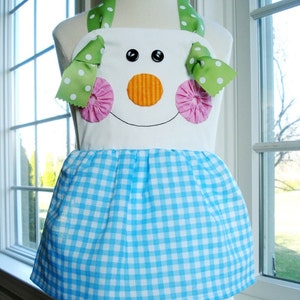 May include: A white apron with a blue and white gingham skirt. The apron has a snowman face with black eyes, an orange nose, and pink cheeks. The apron has green and white polka dot straps with pink and white bows.