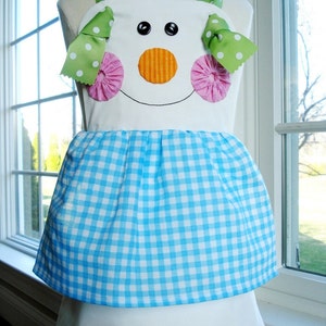 May include: A white apron with a blue and white gingham skirt. The apron has a snowman face with green and white polka dot bows, pink rosettes, and an orange nose.