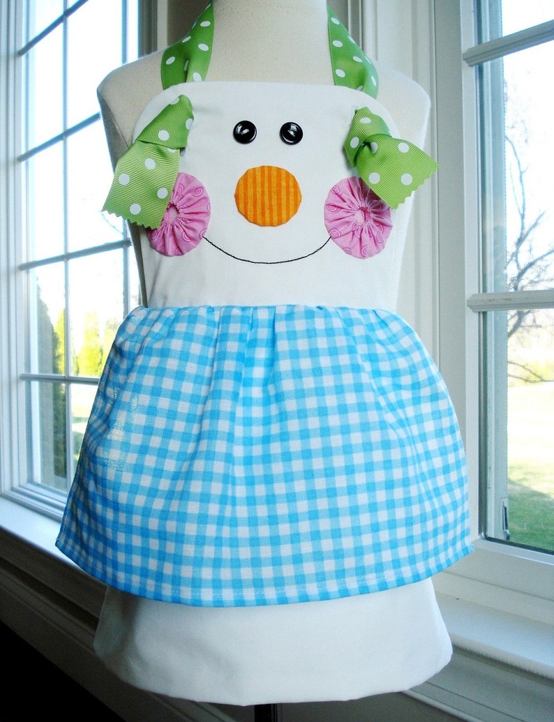 May include: A white apron with a blue and white gingham skirt. The apron has a snowman face with green and white polka dot bows, pink rosettes, and an orange nose.