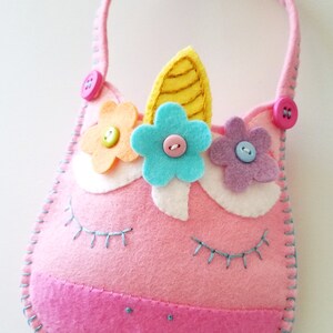Puede incluir: Una bolsa de unicornio de fieltro rosa con un cuerno amarillo, flores de fieltro azul, amarillo y morado, y botones rosas. La bolsa tiene una sonrisa cosida azul y pesta&ntilde;as.
