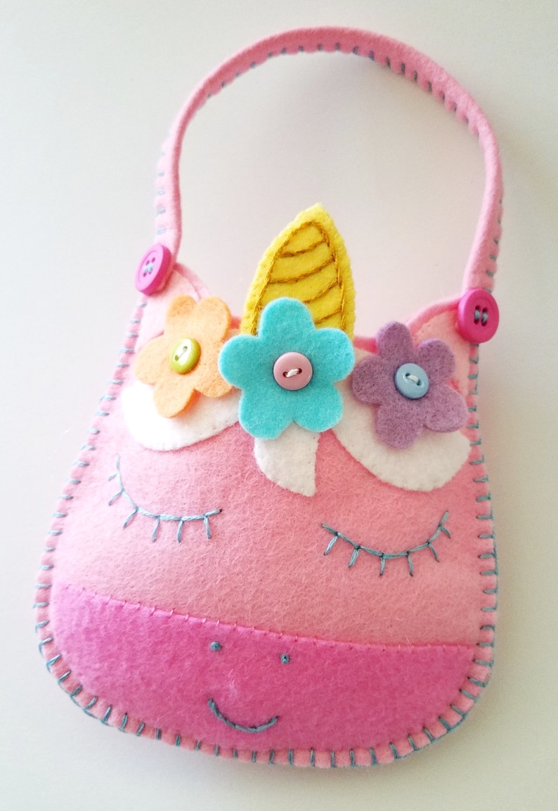 Puede incluir: Una bolsa de unicornio de fieltro rosa con un cuerno amarillo, flores de fieltro azul, amarillo y morado, y botones rosas. La bolsa tiene una sonrisa cosida azul y pesta&ntilde;as.