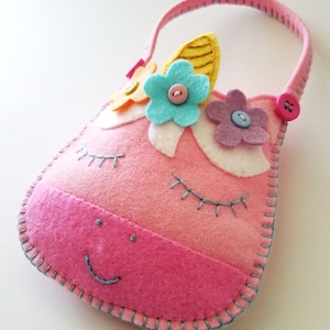 Puede incluir: Bolso de fieltro rosa en forma de unicornio con asa, ojos bordados y boca sonriente. El bolso est&aacute; decorado con flores de fieltro en azul, amarillo y morado, y tiene un cierre de bot&oacute;n.