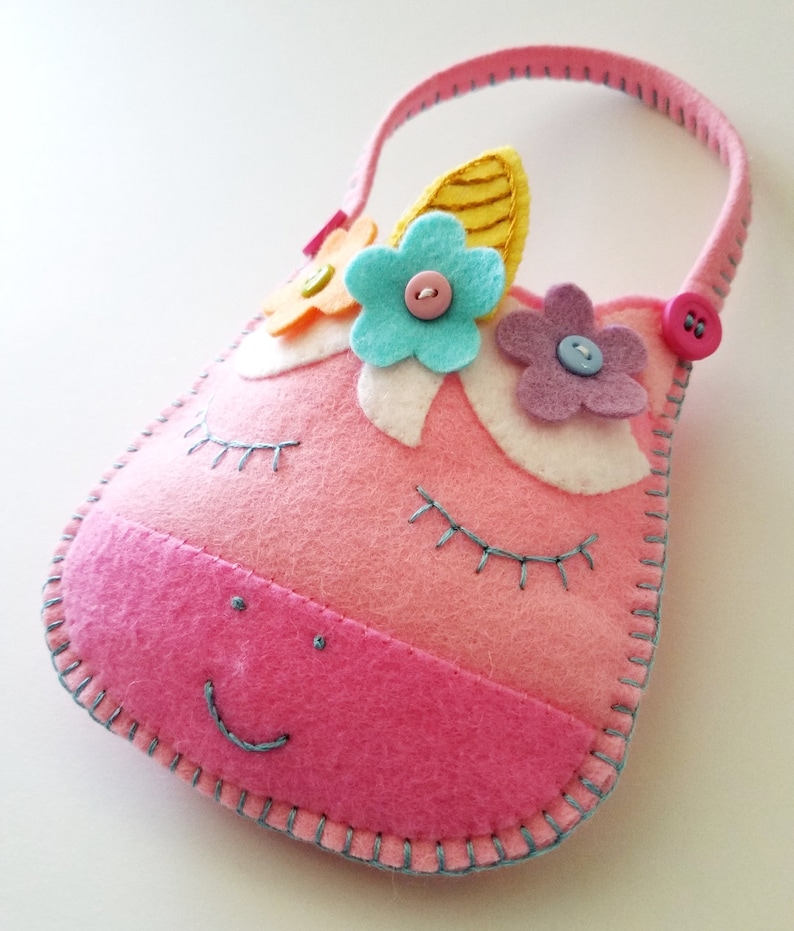 Puede incluir: Bolso de fieltro rosa en forma de unicornio con asa, ojos bordados y boca sonriente. El bolso est&aacute; decorado con flores de fieltro en azul, amarillo y morado, y tiene un cierre de bot&oacute;n.