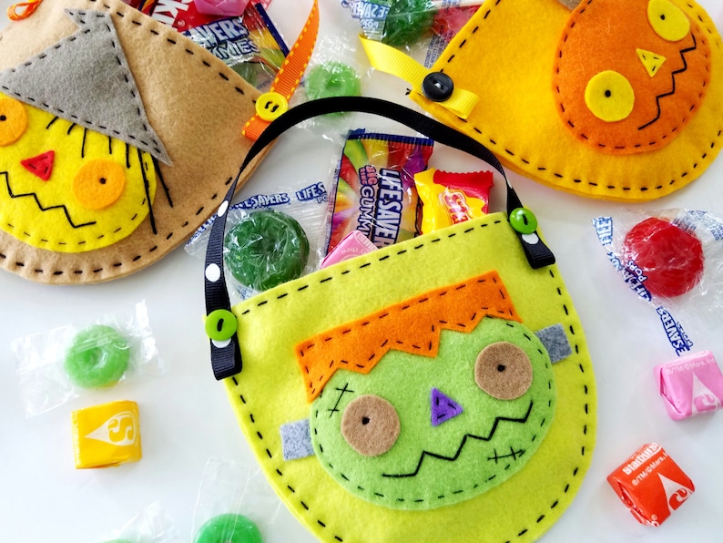 May include: Three felt Halloween trick-or-treat bags with different character designs. The bags are filled with sweets and have black ribbon handles. The characters are a scarecrow, a pumpkin, and a Frankenstein.