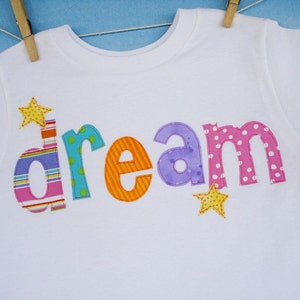 May include: White t-shirt with the word "dream" spelled out in colorful fabric letters. Each letter is a different color and pattern, including stripes, polka dots, and solid colors. There are two yellow stars on the shirt.