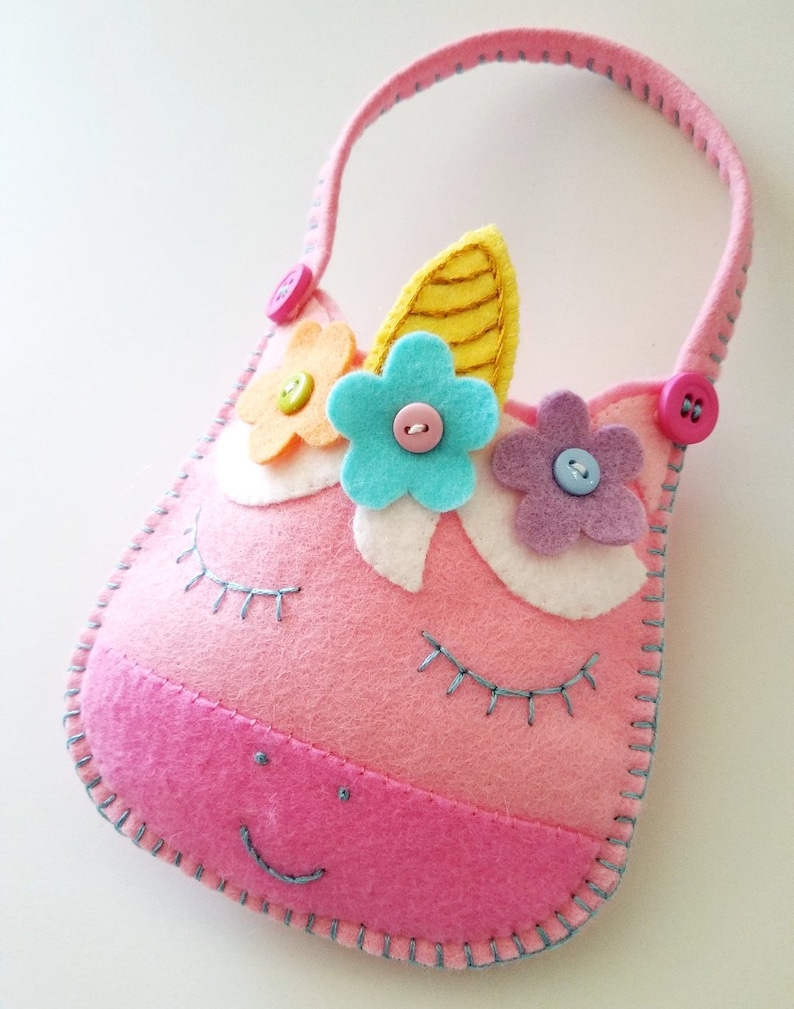 Puede incluir: Una bolsa de fieltro rosa en forma de unicornio con un cuerno amarillo, flores azules y rosas y detalles de botones. La bolsa tiene un asa y una cara sonriente.