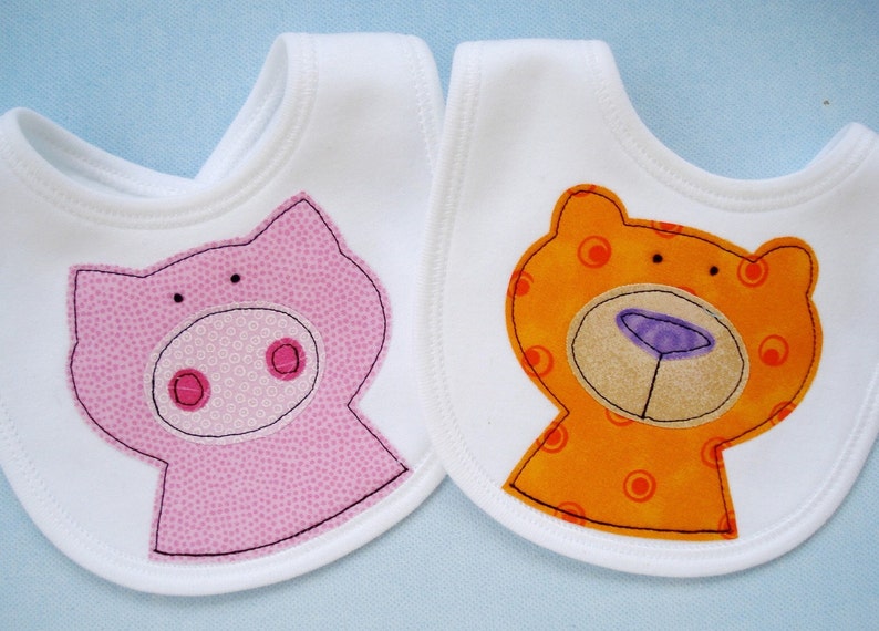 May include: Two white bibs with embroidered animal faces. One bib features a pink pig with a white snout and pink cheeks. The other bib features an orange bear with a brown snout and purple nose.