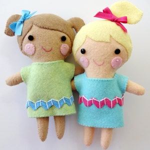 May include: Two felt dolls with embroidered faces and hair. The doll on the left has brown hair, a green dress with a blue trim, and blue bow. The doll on the right has blonde hair, a blue dress with a pink trim, and a pink bow.