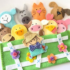 May include: A green felt finger puppet stage with a white picket fence design. The stage features a variety of felt finger puppets including a cow, a duck, a cat, a pig, a horse, a chick, a dog, and a farmer.