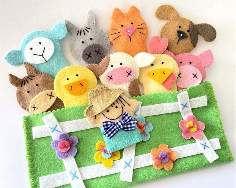 May include: A green felt finger puppet stage with a white picket fence design. The stage features a variety of felt finger puppets including a cow, a duck, a cat, a pig, a horse, a chick, a dog, and a farmer.