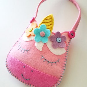 Puede incluir: Bolso de fieltro rosa en forma de unicornio con asa, decorado con flores de fieltro de colores y botones. El bolso tiene ojos bordados y una boca sonriente.