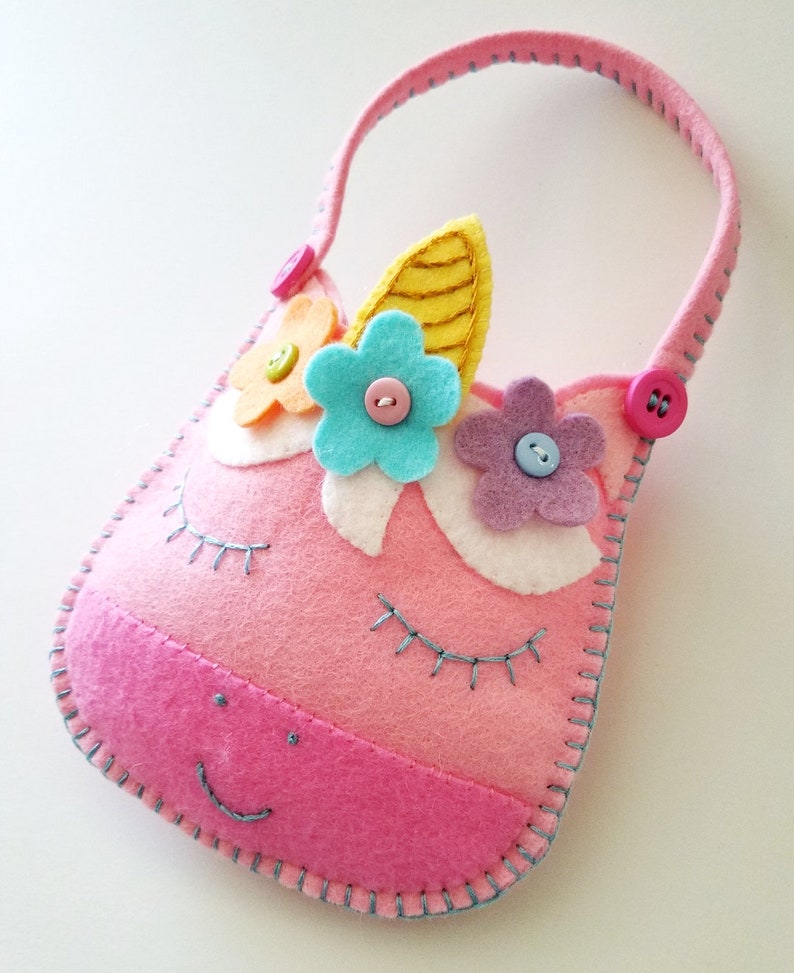 Puede incluir: Bolso de fieltro rosa en forma de unicornio con asa, decorado con flores de fieltro de colores y botones. El bolso tiene ojos bordados y una boca sonriente.