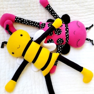 May include: A yellow and black bee plush toy with a smiling face and black eyes. The bee is lying on top of a pink and black ladybug plush toy with a smiling face and closed eyes.