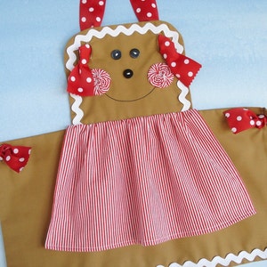 May include: A gingerbread person-shaped apron with red and white polka dot trim and a red and white striped skirt. The apron has a brown face with black eyes and a smile.