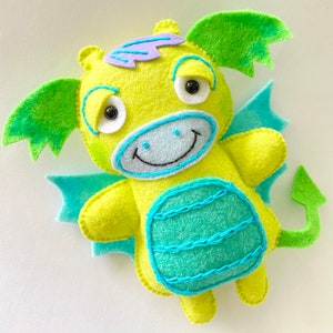 Puede incluir: Un dragón de fieltro verde con alas azules y cola verde. El dragón tiene una cara sonriente con ojos negros y una boca azul claro. El cuerpo del dragón está decorado con un patrón azul claro y verde.