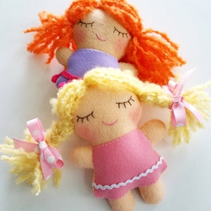 May include: Handmade felt dolls with closed eyes and rosy cheeks. One doll has orange yarn hair, the other has yellow yarn hair tied with pink polka dot ribbons. The dolls wear pink and purple dresses.
