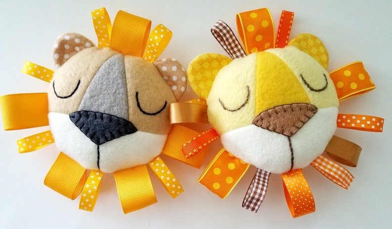 Op de afbeelding: Twee pluche leeuwenkop speelgoed met gele, oranje en witte lint manen. De leeuwen hebben bruine vilt neuzen en gesloten ogen.