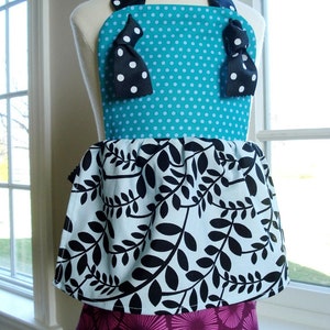 May include: A turquoise and white polka dot apron with black bows and a black and white leaf print skirt. The apron has a pink and white floral print bottom layer.