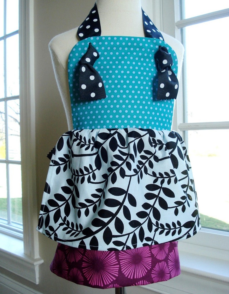 May include: A turquoise and white polka dot apron with black bows and a black and white leaf print skirt. The apron has a pink and white floral print bottom layer.