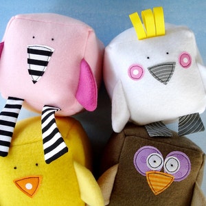 Puede incluir: Tres bloques de juguetes de peluche con forma de pájaros. El primer bloque es rosa con rayas blancas y negras, el segundo es blanco con una cresta amarilla y mejillas rosas, y el tercero es marrón con ojos morados y un pico naranja.