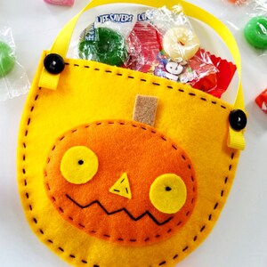 May include: A yellow felt pumpkin trick-or-treat bag with a black stitched outline and a felt orange pumpkin face with yellow eyes and a nose. The bag has a yellow ribbon handle with black buttons.