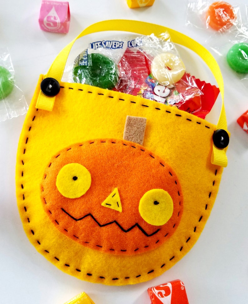 May include: A yellow felt pumpkin trick-or-treat bag with a black stitched outline and a felt orange pumpkin face with yellow eyes and a nose. The bag has a yellow ribbon handle with black buttons.