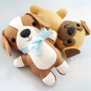 May include: Two felt stuffed toy dogs. One is brown and white with a blue bow and the other is tan with brown ears and a brown face.