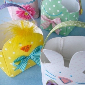 May include: Four small Easter baskets made of fabric with different animal designs. The baskets are decorated with polka dots, ribbons, and embroidered details. The animals include a chick, a bunny, a frog, and a pink flower.