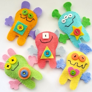 Puede incluir: Cinco marionetas de dedo de monstruos de fieltro con diferentes colores, formas y ojos de botón. Las marionetas son verdes, azules, amarillas, naranjas y rosas.