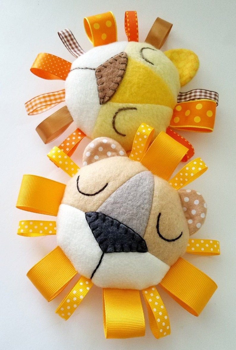 Op de afbeelding: Twee pluche speelgoedjes in de vorm van een leeuwenkop met gele, oranje en witte viltgezichten. Elke leeuw heeft een manen van geel lint met witte stippen.