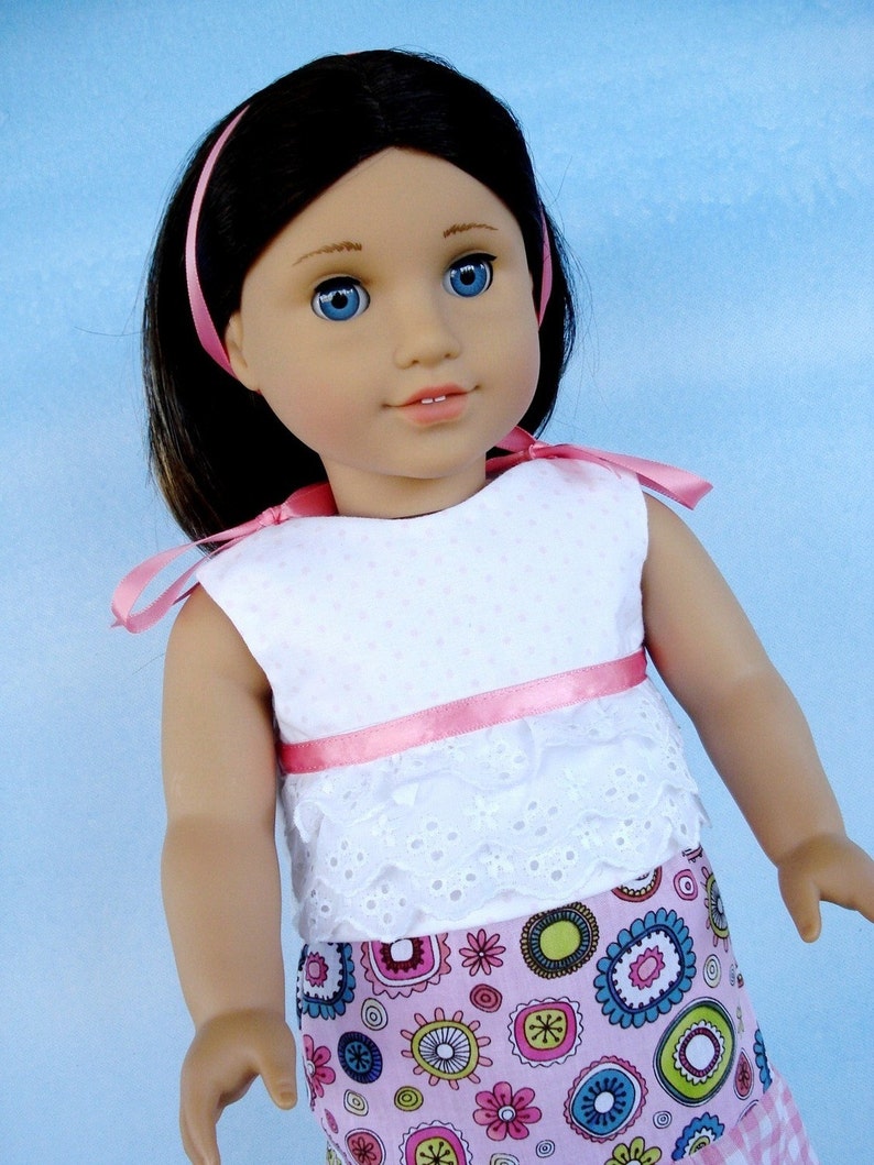 May include: A doll with long brown hair wearing a white and pink dress with a pink ribbon headband and a colorful floral print skirt.