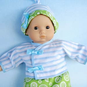 Puede incluir: Una muñeca bebé con una camisa a rayas azules y blancas con lazos azules, un gorro verde y blanco con círculos verdes y pantalones verdes y blancos con círculos verdes.