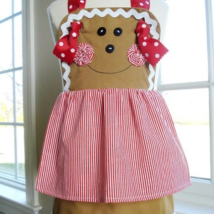 May include: A gingerbread person-shaped apron with red and white striped fabric and red and white polka dot ribbon accents. The apron has a white scalloped trim.