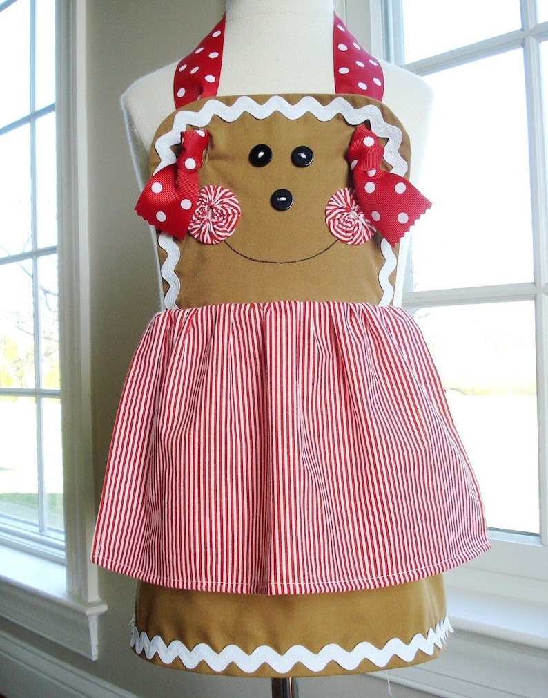 May include: A gingerbread person-shaped apron with red and white striped fabric and red and white polka dot ribbon accents. The apron has a white scalloped trim.