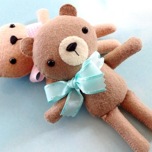 May include: Two brown felt teddy bears, one with a light blue bow and the other with a pink and white gingham bow. The bears are lying on a light blue background.