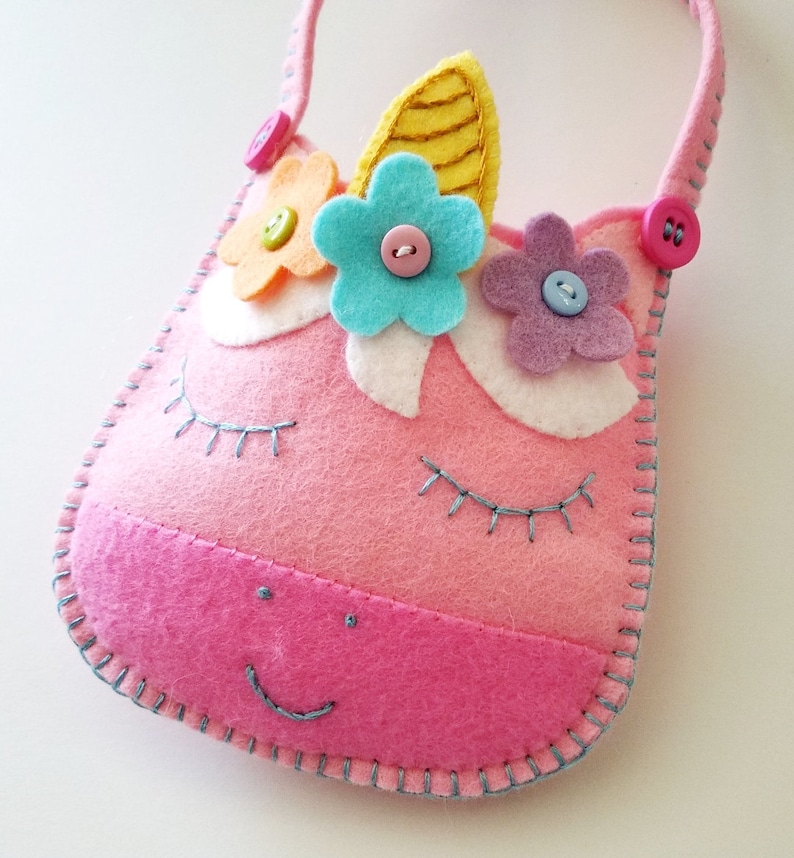 Puede incluir: Un bolso de unicornio de fieltro rosa con un cuerno amarillo, flores azules, rosas y amarillas, y un bot&oacute;n rosa. El bolso tiene una cara de fieltro blanco con pesta&ntilde;as bordadas y una sonrisa de fieltro rosa.