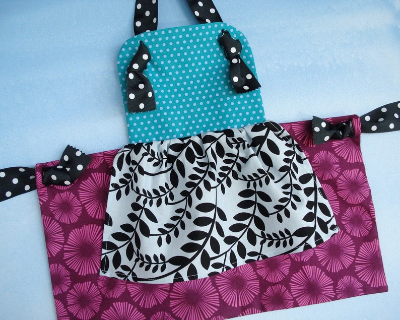 May include: A turquoise and white polka dot fabric apron with black ribbon ties and a black and white leaf print ruffle. The apron has a pink and white floral print bottom.