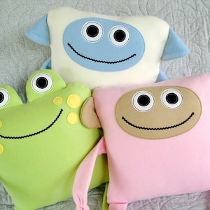 Puede incluir: Tres juguetes de peluche con caras sonrientes. Una rana verde, una oveja azul y blanca y un mono rosa.