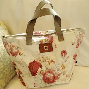 May include: A large, off-white canvas tote bag with a floral print featuring red roses, white daisies, and green leaves. The bag has brown leather handles and a zipper closure.